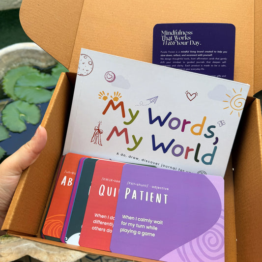 A hand holding an open cardboard box containing a journaling kit. The kit includes a white cover card titled 'My Words, My World,' a purple 'Mindfulness That Works Today Your Day,' and several colorful prompt cards in orange, teal, gray, and purple.